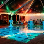 Experience a magical nighttime event in North Georgia with T3 Event Rentals. Our stunning setup features a transparent tent cover, beautifully illuminated by vibrant lights that create an enchanting atmosphere. Inside the tent, round tables adorned with white tablecloths are elegantly arranged on a reflective floor, enhancing the elegant ambiance of your special occasion. Remember, all items shown here are rentals from T3 Event Rentals – serving exquisite events throughout North Georgia.