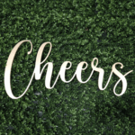 The image showcases a stylish cursive wooden cutout rental sign, spelling Cheers, elegantly placed against lush green foliage. Perfect for enhancing your events in Dawsonville, this piece is available exclusively through T3 Event Rentals.
