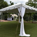 A sophisticated white outdoor tent featuring elegant drapery and pole covers is set up on a lush grassy area in Forsyth County. In the background, you can spot a DJ booth complete with speakers, decorative string lights, and a few attendees enjoying the scene. The entire setup is surrounded by beautiful trees and greenery. All these items are provided by T3 Event Rentals, specializing in exceptional event experiences in Forsyth County and beyond.