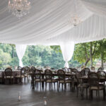 Experience an exquisite outdoor event setting in Dawsonville with T3 Event Rentals. Our expansive tented venue boasts elegant chandeliers, draped white ceilings, and ambient strings of lights. Rustic wooden chairs encircle tables offering stunning views of lush greenery and a serene body of water. Ideal for creating unforgettable moments, T3 Event Rentals brings your vision to life in the heart of Dawsonville.
