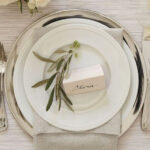 Experience elegance at your next Atlanta event with T3 Event Rentals stunning tableware. Featuring a place setting complete with a white plate on a sophisticated silver charger, complemented by a folded beige napkin beneath. A personalized touch is added with a small white card bearing the name Olivia, highlighted by delicate sprigs of greenery. Complete your table decor with elegant silverware arranged beside the plate—all available for rent through T3 Event Rentals, where we bring style and grace to every occasion in Atlanta.