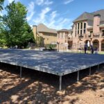 Discover the perfect event setting with T3 Event Rentals in Dawsonville. Our expansive outdoor platform, elevated on sturdy metal supports, provides an ideal venue backdrop near charming brick and yellow buildings under a picturesque blue sky adorned with clouds. Surrounding trees and a thoughtfully placed fence enhance the ambiance, ensuring your occasion is both memorable and picturesque.