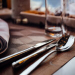 Close-up of a dining table setup, highlighting a shiny fork, knife, and spoon laid on a brown woven placemat in Forsyth County. A folded napkin sits to the left, accompanied by glasses in a softly blurred backdrop that hints at a cozy dining ambiance. All items are rentals from T3 Event Rentals, serving beautiful Forsyth County with elegance and style.