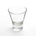 A clear, empty shot glass with a slightly tapered design is showcased against a plain white background, capturing the aesthetic of an Optic White Wine Glass from a 25 rack. Its thick base emphasizes clean, minimalist lines that resemble those found in modern wine glass racks. Available for rent in Atlanta through T3 Event Rentals, renowned for supplying quality event items throughout the area.