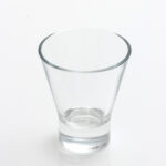 A clear, empty shot glass with a tapered shape sits gracefully on the Optic White Wine Glass (25 rack), casting soft reflections on the solid white surface below. This elegant combination, available for rent from T3 Event Rentals, adds a touch of sophistication to any event in Atlanta.