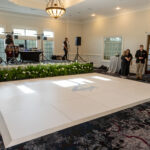 A beautifully decorated indoor event space in Dahlonega features a stunning White Dance Floor with a blue emblem available from T3 Event Rentals. Lush greenery accents and a stage equipped with professional musical equipment create the perfect backdrop for your next celebration. A few formally dressed guests mingle in this elegant setting, showcasing the transformative event rental services offered by T3 Event Rentals throughout Dahlonega and surrounding areas. Please note, all items pictured are rentals only—nothing is for sale.