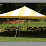 A vibrant 20 Rope and Pole Tent with yellow and white stripes is elegantly set up on grass in Dawsonville. Inside, tables and chairs are arranged in a circular layout, creating a welcoming atmosphere. The scenic backdrop includes trees and a picturesque dirt path. All items are rentals provided by T3 Event Rentals. Based in Alpharetta, T3 Event Rentals proudly serves the Dawsonville area with top-quality event essentials.