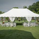 A 20 Rope And Pole Tent from T3 Event Rentals stands on a grassy field in Atlanta, surrounded by rows of elegant white chairs. Beautiful hanging flower baskets decorate the tents corners, while nearby trees and an overcast sky contribute to the serene ambiance.