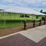 A classic 20 Rope And Pole Tent is beautifully erected on a lush lawn under a cloudy sky in Forsyth County. Nearby, someone in a red shirt adds life to the scene at the tents edge. In front of this picturesque setting, enjoy the wooden deck with patio furniture that perfectly complements the atmosphere. In the distance, admire the serene trees and rolling hills for a complete outdoor experience. All items featured are available for rent from T3 Event Rentals—your trusted event partner serving Forsyth County and beyond. Choose T3 Event Rentals for all your event needs in North Georgia!.