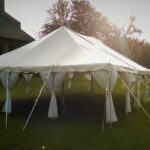 A 20 Rope And Pole Tent, courtesy of T3 Event Rentals, is elegantly set up outdoors on the lush grass of Forsyth County. Anchored securely with stakes, the tent glows beautifully in the sunlight. To the left, a stone wall building peeks into view while majestic trees form a picturesque background. Although these are premium rentals from T3 Event Rentals and not for sale, they transform any location into an extraordinary setting.