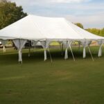 A 20 Rope and Pole Tent from T3 Event Rentals is beautifully set up in a grassy field surrounded by trees in Dawsonville. The tent features supporting poles and side curtains tied back for an open-air feel, blending seamlessly with the partly cloudy sky above. Perfect for any event in Dawsonville, this rental from T3 Event Rentals offers both elegance and practicality.