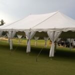 A 20 Rope and Pole Tent, available from T3 Event Rentals, is elegantly set up in Dawsonville on a grassy field with draped entrances and supported by ropes and poles. Nearby, a truck is parked while trees stand tall in the background under a partly cloudy sky. All items you see are rentals from T3 Event Rentals, perfect for your next event in Dawsonville.