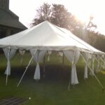 A beautiful 20 Rope and Pole Tent from T3 Event Rentals is elegantly positioned on a lush grassy lawn near a stone-walled building in Forsyth County. The bright sun creates long, dramatic shadows, while distant trees and a partially visible vehicle add to the charming ambiance of this North Georgia location. All items featured are rentals from T3 Event Rentals.