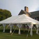 A 20 Rope And Pole Tent in white is elegantly set up on a manicured lawn with open sides and multiple supporting poles, providing a perfect rental solution by T3 Event Rentals. In the heart of Atlanta, a stone building with a steep roof creates an impressive backdrop, while an individual stands by the tent on the left. Remember, T3 Event Rentals specializes in transforming spaces across Atlanta for your special occasions.