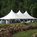 A stunning 20 rope and pole tent with three peaks is set up on a lush green lawn in Atlanta, encircled by magnificent trees. Underneath the tent, tables draped in white linens and adorned with pink decorations invite guests to enjoy the gathering. The picturesque scene is completed by a charming flower bed with rocks in the foreground. All items showcased are rentals from T3 Event Rentals, providing top-notch service for events throughout Atlanta.