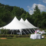 A stunning 20-foot Rope and Pole Tent with three peaks is elegantly set up on a grassy area encircled by trees in Dawsonville. Nearby, under the partly cloudy sky, a round table draped with a white cloth showcases various rental items for an event. All items are provided by T3 Event Rentals. Although based in Alpharetta, T3 Event Rentals frequently services events across Dawsonville and beyond.
