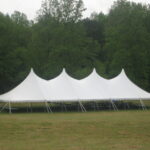 A 20 Rope and Pole Tent with peaked roofs stands elegantly on a verdant field in Dahlonega, surrounded by lush trees. The clear sky enhances the scene, perfectly showcasing the sophistication of this white event tent rental from T3 Event Rentals. All items are available for rent.