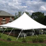 T3 Event Rentals brings to life a stunning outdoor scene with a 20 Rope And Pole Tent set up on a lush lawn near a charming brick house in Dawsonville. Inside, round tables and chairs await guests for an upcoming gathering. Majestic trees provide a natural backdrop under the partly cloudy sky, enhancing this beautiful setting. If youre planning an event in Dawsonville or nearby areas, consider T3 Event Rentals for all your party rental needs.