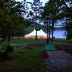 Discover the charm of a 20 Rope And Pole Tent glowing warmly by a lakes edge in Forsyth County. Surrounded by trees and connected by a stone path, this enchanting setup invites guests to enjoy the early evening ambiance. T3 Event Rentals offers this exquisite rental experience, ensuring your event is unforgettable in Forsyth County. Explore our services and let T3 Event Rentals transform your gathering into something magical amidst natures beauty.