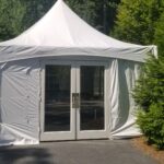 T3 Event Rentals offers a 10 frame tent featuring a pointed roof and glass double doors, perfect for your next event in Dawsonville. Situated on a paved area surrounded by lush greenery, this stunning tent shines under the bright sunlight. The structure is equipped with a white ventilation tube for added comfort. Remember, all items are rentals from T3 Event Rentals.