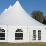 A majestic white event tent with pointed peaks and arched windows, surrounded by sidewalls, is set up on the lush grassy grounds in Dawsonville. The entrance features elegant double doors and a charming wooden building can be seen nearby under the clear blue sky. All items featured are rental services provided by T3 Event Rentals.