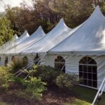 A picturesque row of white event tents with pointed tops and arched windows from T3 Event Rentals stands elegantly on a grassy area in Forsyth County. The sunny skies cast charming shadows as these rental tents are complemented by sidewalls and surrounded by lush shrubs and trees.