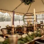 Experience the enchanting ambiance of a beautifully adorned tented space in Atlanta with sidewalls that include a wooden table, elegant glassware, tall candles, and a round metal shelf of bottles. The addition of greenery and white flowers enhances the natural feel, while soft lighting creates a warm atmosphere. This stunning setup features rental items from T3 Event Rentals, your go-to source for event rentals in the area.