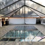 Transform your next event with T3 Event Rentals stunning clear tent setup in Atlanta. Featuring a unique glass-like floor and elegant wooden flooring along the edges, this tent can upgrade any location ambiance. Delicate string lights outline the perimeter, creating a magical atmosphere. With seating visible on the left and two people welcoming guests near the entrance, its perfect for all occasions. Plus, sidewalls offer added privacy and protection for events above pools or other special settings. All these elements are available through T3 Event Rentals, ensuring your Atlanta event is extraordinary without purchasing any items.