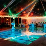 Experience the magic of a festive outdoor event in Forsyth County, where a clear tent with sidewalls houses tables adorned with white cloths and multicolored lights from T3 Event Rentals. The elegant atmosphere is enhanced by a glowing pool, surrounded by chic chairs and decor. T3 Event Rentals specializes in creating unforgettable events in Forsyth County with top-quality rental items.