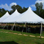 A picturesque 20 Rope and Pole Tent with peaked tops is elegantly set on grass under a partly cloudy blue sky in Forsyth County. Under the tent, tables and chairs are arranged perfectly, creating an inviting atmosphere. The surrounding trees add to the natural charm of this Forsyth County location. All items, including this beautiful setup, are rentals provided by T3 Event Rentals. Based in Alpharetta, T3 Event Rentals proudly serves Forsyth County and other neighboring areas with top-notch event rental services.