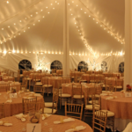An exquisite wedding reception takes place under a spacious 20 Rope and Pole Tent in Dahlonega, featuring elegant round tables with pink tablecloths and beige chairs, all provided by T3 Event Rentals. The ambiance is enhanced by string lights overhead, casting a welcoming glow while table numbers are prominently displayed for guests convenience.
