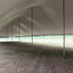 A magnificent 20 Rope And Pole Tent by T3 Event Rentals graces a grassy field in Forsyth County. The tents elegant white design features multiple peaks, supported by sturdy metal poles, providing partial shade as sunlight filters through. Perfect for your next event, trust T3 Event Rentals to deliver top-quality rentals in the Forsyth County area.