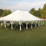 Our 20 Rope And Pole Tent, available for rent from T3 Event Rentals, is elegantly set up on a grassy field amidst lush greenery and trees in Forsyth County. This large white event tent features open sides that offer breathtaking views of the surrounding landscape and sky beyond. Perfect for your next gathering, T3 Event Rentals ensures a seamless experience with our premium rental options.