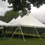 Under an overcast sky in Atlanta, a person stands on a ladder fine-tuning the setup of a 20 Rope and Pole Tent. The large white rental tents from T3 Event Rentals dot the grassy outdoor area among trees, braving the gentle breeze and hinting at impending rain.