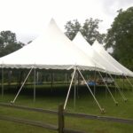 A 20 Rope And Pole Tent with striking pointed peaks is beautifully set up on a lush green lawn in Atlanta, creating an idyllic scene surrounded by trees and a wooden fence in the foreground. The overcast sky complements the serene ambiance of this eye-catching tent rental arrangement. For those seeking event rentals in Atlanta, T3 Event Rentals is your go-to company for creating memorable experiences.