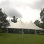 In a grassy field beneath the cloudy skies of Forsyth County, a spacious white 20 Rope And Pole Tent from T3 Event Rentals stands elegantly among the trees. A small gathering of people enjoys the ambiance under the tent, while a white truck idles nearby, perfectly integrated into the scene by the tent’s 20-foot rope.
