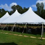 T3 Event Rentals creates the perfect setting in Forsyth County with a 20 Rope And Pole Tent, featuring three elegant peaked tops, set against a serene grassy landscape surrounded by trees. Under this beautiful tent are tables and chairs available for rent, offering a comfortable gathering space. A charming stone path leads guests to the entrance, all under a partly cloudy sky that accentuates the idyllic atmosphere.