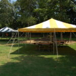 Image 1: In Dahlonega, a picturesque scene unfolds with two outdoor tents set up on a lush grassy field. A vibrant yellow and white striped 20 Rope And Pole Tent from T3 Event Rentals stands prominently, offering an inviting space filled with arranged tables and chairs. Tall trees frame the background underneath a clear sky, creating an ideal setting for any occasion. Image 2: Discover the charm of North Georgia with T3 Event Rentals exquisite setups. On this serene landscape, a striking black and white striped tent complements the natural beauty of North Georgia perfectly. Whether planning events in Dahlonega or beyond, rely on T3s rental expertise to craft memorable experiences.