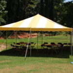 A vibrant 20 Rope and Pole Tent with striking yellow and white stripes is elegantly set up outdoors on lush grass in Atlanta. Beneath the tent, several round tables paired with chairs are arranged comfortably, creating an inviting setting against a scenic backdrop of trees and a small dirt area. All items pictured are available for rent through T3 Event Rentals, ensuring your event is both stylish and seamless. Located in Alpharetta, T3 Event Rentals proudly serves the Atlanta area for all your special occasion needs.