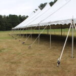 In a picturesque grassy field in Dahlonega, a 20 Rope and Pole Tent from T3 Event Rentals elegantly stretches across the landscape. Anchored by several poles and secured with ropes, it creates a charming scene amidst the lush surroundings. The classic setup is beautifully completed by the silhouette of trees against a cloudy sky. Whether youre planning an event in Dahlonega or elsewhere, T3 Event Rentals provides all your rental needs to make it unforgettable.