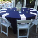 Plan your outdoor event in Dawsonville with an elegant round table setup by T3 Event Rentals. Featuring a striking dark tablecloth complemented by pink napkins and pristine white chairs, this setting is perfect for any gathering. A stunning flower-filled decorative birdcage serves as the centerpiece, gracefully encircled by small candles on the wooden deck. Ideal for events throughout Dawsonville, T3 Event Rentals provides top-notch rental services to bring your vision to life.