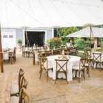 Experience the enchanting outdoor wedding reception in North Georgia with T3 Event Rentals. Our setup features round tables draped in elegant white tablecloths, perfectly paired with wooden chairs and simple floral centerpieces, all nestled under a canopy. In the foreground, enjoy the ambiance of a candle-adorned wooden rectangular table set amidst the lush garden setting. T3 Event Rentals brings this refined touch to your North Georgia celebrations with our exquisite rentals.