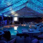 Experience an enchanting evening event in Atlanta, hosted under a spacious white tent adorned with captivating blue lights. Surrounding the sparkling pool are elegantly set round tables, each covered with pristine white cloths and accented by black sashes. Overhead, blue string lights from T3 Event Rentals create dazzling patterns against the tents ceiling. All event furnishings are high-quality rentals provided by T3 Event Rentals.