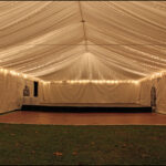Imagine a delightful setting in Atlanta and North Georgia, where a spacious white rental tent with an elegantly draped ceiling and twinkling string lights creates the perfect ambiance. Nestled within Forsyth County and easily accessible from Dawsonville, Dahlonega, and Marietta, this venue features an Indoor Stages small stage at the back. The scene is completed by a wooden dance floor ready for guests to enjoy, two tall round tables flanking either side, and scattered leaves adding charm to the grassy ground beneath.