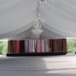 Introducing our Indoor Stages – a premium event space for rental nestled in the heart of North Georgia. Featuring a spacious white tent interior adorned with crystal chandeliers and gray carpeted flooring, it creates an exquisite setting for elegant events. The maroon-white curtains at the back add a touch of sophistication, while the side openings offer picturesque views of surrounding trees. Perfect for hosting memorable experiences in Atlanta, Forsyth County, Dawsonville, Dahlonega, and Marietta, our venue provides an unparalleled ambiance that elevates any occasion.