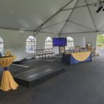 Inside a rental white tent featuring arched windows, an Indoor Stage is set up next to a high table decorated with a yellow tablecloth in Atlanta, North Georgia. Nearby, a larger table draped in blue and yellow showcases framed photos, ideal for events in Forsyth County, Dawsonville, Dahlonega, and Marietta. In the backdrop, chairs and a screen add to the elegant setup commonly seen across these scenic regions of North Georgia.