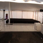 A spacious white tent in the heart of North Georgia houses an ideal indoor stage for event rentals, perfect for venues in Atlanta, Forsyth County, Dawsonville, Dahlonega, and Marietta. This elegant space features beautifully tiled flooring surrounded by chic white cube-shaped seating and tables. In the background, a banner and potted plants add a refreshing touch of greenery to your event setting.
