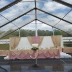 Experience the sophistication of an elegant wedding reception setup in the heart of North Georgia, featuring an indoor stage rental with a pristine clear tent. Ideal for locations such as Atlanta, Forsyth County, Dawsonville, Dahlonega, and Marietta, this venue includes a stunning white and gold draped backdrop. The head table is beautifully adorned with pink and gold fabrics alongside exquisite floral arrangements. Located outdoors, the transparent tent walls perfectly frame the surrounding trees for guests to enjoy amidst these picturesque areas.