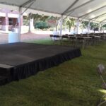 A spacious outdoor rental tent is set up on grass in North Georgia, showcasing a small stage complete with steps and a black skirt. Inside, long rows of tables and chairs are arranged for an event against a picturesque backdrop of trees and brick architecture. Whether youre hosting in Atlanta, Forsyth County, Dawsonville, Dahlonega, or Marietta, our indoor stages enhance any occasion.