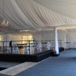 Experience the elegance of The Indoor Stages rental event tent in North Georgia, featuring a high white draped ceiling adorned with chandeliers. Located conveniently for those in Atlanta, Forsyth County, Dawsonville, Dahlonega, and Marietta, this sophisticated venue is enhanced by white chairs and tables set against a chic gray carpet floor. With its minimal decor allowing for versatile arrangements, its the perfect space to host your next event in stunning North Georgia.